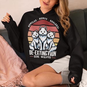 May include: Black hooded sweatshirt with a retro sunset graphic featuring three white dire wolves and the text "Romulus - Remus - Khaleesi De-Extinction - Dire Wolves".