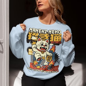 May include: Pale blue sweatshirt featuring a cartoon Maneki-Neko cat in a city scene with Japanese text. The design includes the phrases "MANEKI-NEKO ATTACK" and "MAY LUCK BE WITH YOU!"