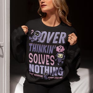 Puede incluir: Sudadera negra con el texto "OVER THINKIN' SOLVES NOTHING" en morado y rosa, con ilustraciones de robots. La frase "STOP WORRYING START CREATING" está impresa debajo del texto principal.