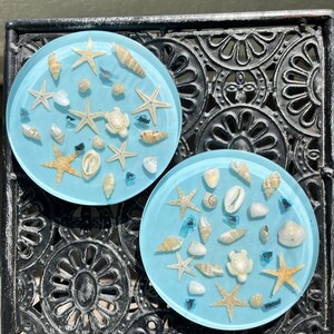 Puede incluir: Dos posavasos redondos de color azul claro con incrustaciones de conchas marinas, estrellas de mar y pequeños trozos de vidrio azul. Los posavasos están sobre una mesa decorativa de metal negro, creando un tema costero.