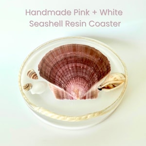 Puede incluir: Un posavasos redondo de resina hecho a mano con una gran concha rosa como pieza central, rodeada de conchas más pequeñas. El posavasos tiene un acabado transparente y brillante con un borde en forma de cuerda. El texto en la parte superior dice "Handmade Pink + White Seashell Resin Coaster."