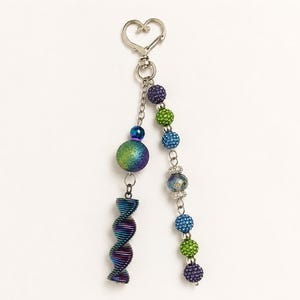 Puede incluir: Un llavero de tono plateado con un cierre en forma de corazón unido a dos cadenas colgantes. Una cadena sostiene una esfera grande, iridiscente y texturizada y un colgante metálico en espiral. La otra cadena presenta una serie de pequeñas cuentas texturizadas de colores en tonos morado, verde y azul, intercaladas con cuentas transparentes facetadas.
