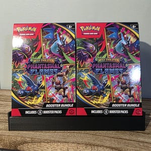 May include: Two Pokémon TCG booster bundles on a black display stand. Each bundle features the text "Mega Evolution Phantasmal Flames" and includes 6 booster packs. The packaging has a red and black color scheme with various Pokémon characters.