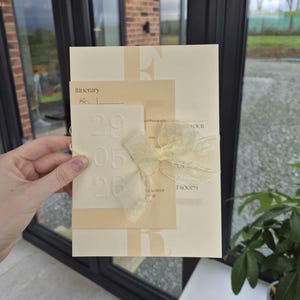 May include: A cream-colored wedding invitation suite with a light yellow ribbon. The invitation includes the word "Itinerary" and the date "29 05 26" embossed on a card. The suite is held by a hand.