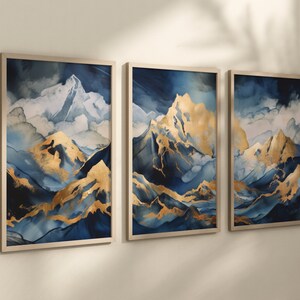 Op de afbeelding: Een set van drie ingelijste abstracte landschapschilderijen met een bergketen in goud- en blauwtinten. De schilderijen zijn ingelijst in een licht houten lijst.
