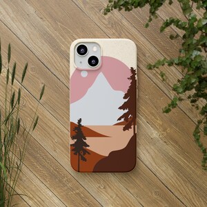 May include: A phone case featuring a minimalist landscape design. The case has a beige background with a pink sun, a white mountain, and brown trees and hills. The phone case is on a wooden surface.