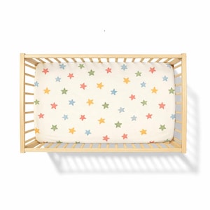 May include: A light-colored crib with a fitted sheet featuring a pattern of colorful stars. The stars are in shades of red, yellow, green, and blue, set against a cream background. The crib is made of light-colored wood.
