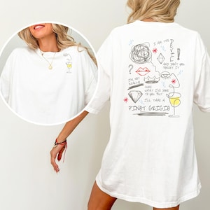 May include: White t-shirt with a minimalist wine glass design on the front and a playful illustration on the back. The back graphic includes the text "I AM THE DEVIL" and "I'LL TAKE A PINOT GRIGIO."