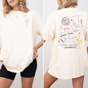 May include: Cream-colored oversized t-shirt with a line drawing of a face and a wine glass on the front. The back features a sketched design with text including "I'll take a Pinot Grigio."