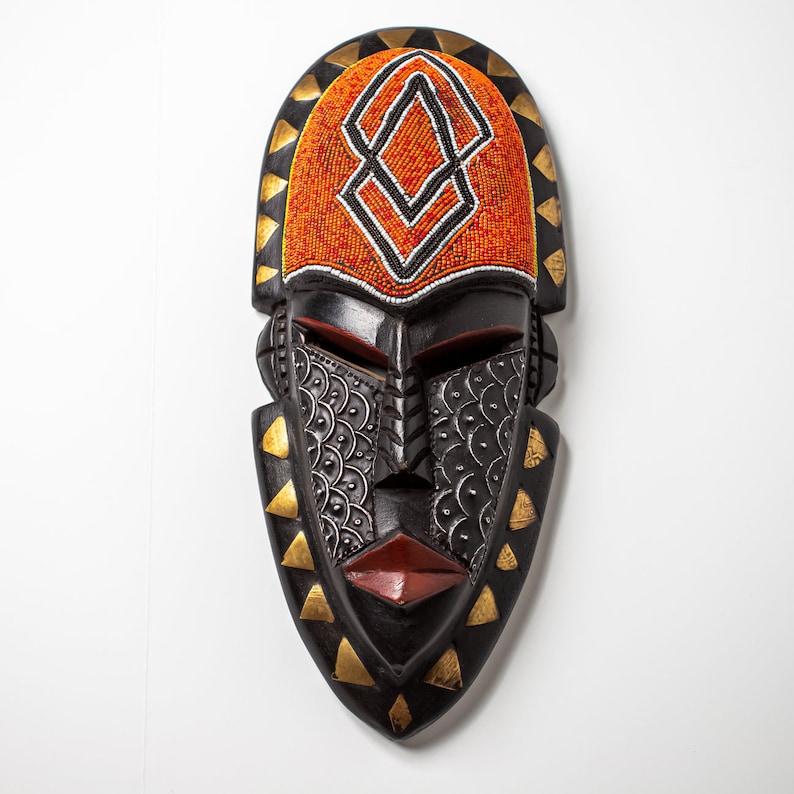 May include: A black wooden mask with a geometric design. The mask has a red and orange beaded pattern on the forehead and gold accents around the edges.
