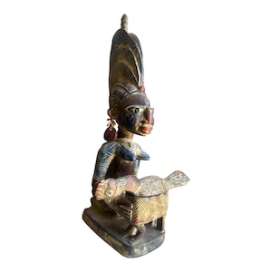 May include: A hand-carved wooden sculpture featuring a figure with an elaborate, tall headdress and blue patterned arms, holding a bird-shaped vessel. The figure has dark skin, painted facial features, and a small red accent on the forehead.