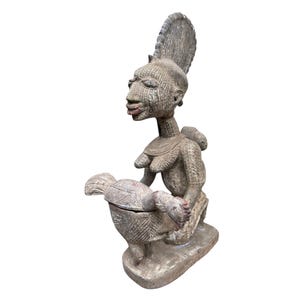 Puede incluir: Escultura de madera tallada gris de una figura sentada con un niño en la espalda. La escultura presenta detalles tallados, incluyendo rasgos faciales, ropa y un recipiente sostenido en el regazo de la figura. La figura tiene un tocado alto.