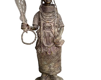 May include: A bronze-toned statue of a figure wearing a detailed headdress and ornate clothing. The figure holds a stylized feather in one hand and a ring-like object in the other. The statue has a textured surface and stands on a round base.