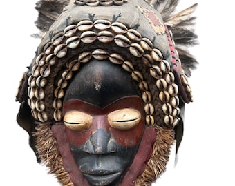 May include: A wooden mask with a black face, red lips, and closed eyes. The mask is decorated with shells, feathers, and a red and brown headdress.