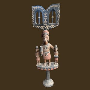 May include: A carved wooden sculpture featuring a central figure holding a rectangular object, flanked by two smaller figures. The sculpture has a decorative top with a blue and white design, and stands on a pedestal. The color palette includes shades of brown, red, and blue.