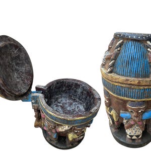Puede incluir: Una caja de madera con una tapa con bisagras decorada con figuras talladas y pintada en azul, dorado y marrón. La caja está abierta, revelando el interior.