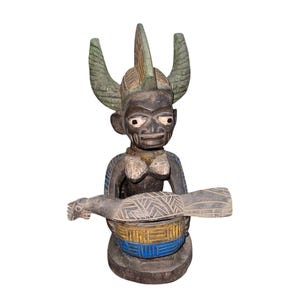 May include: A carved wooden statue featuring a figure with a horned headdress and a bird-like object. The statue is primarily dark brown with accents of green, blue, and yellow. The figure has a detailed face and is holding the bird-like object.