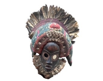 May include: A wooden mask with a dark brown face, large eyes, and a wide mouth. The mask is decorated with colorful fabric, feathers, and cowrie shells.