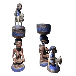 May include: Two carved wooden sculptures with blue and brown accents. One sculpture features a figure atop a box, another figure, and a base. The other sculpture features a figure carrying a bowl, a child, and a figure riding an animal.