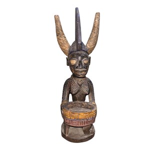 May include: A carved wooden statue of a figure with a horned headdress and a bowl-shaped base. The statue is primarily brown with gold and red accents on the bowl. The figure has detailed facial features.
