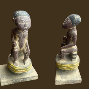 May include: Two wooden sculptures of seated figures. The figures have dark skin and wear head coverings. They are seated on a round base with yellow and green accents, atop a rectangular base. The sculptures are weathered.