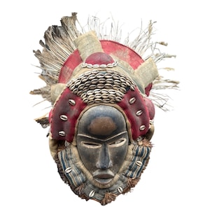 Puede incluir: Una máscara de madera con rostro humano, decorada con conchas de cauri, tela roja y fibras deshilachadas. La máscara tiene un rostro marrón oscuro con una expresión seria y es una pieza de arte tradicional africana.