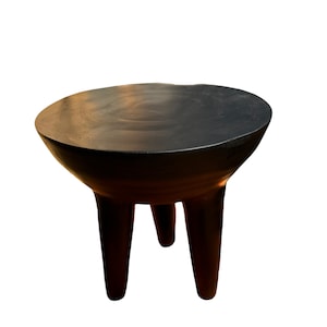 Puede incluir: Un taburete de madera negro con una parte superior redonda y tres patas. El taburete está hecho de madera oscura y tiene un acabado liso.