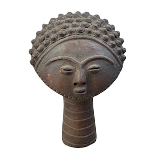 May include: A terracotta sculpture of a stylized human head with a textured, circular head and a cylindrical neck. The head has a simple, stylized face with closed eyes, a small nose, and a slightly open mouth.