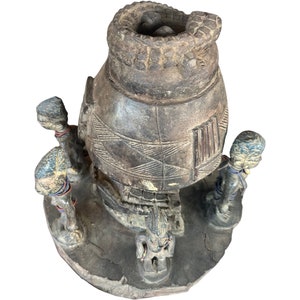 Könnte beinhalten: Eine Holzskulptur, die ein traditionelles afrikanisches Dorf darstellt, mit einer großen, runden Hütte in der Mitte und kleineren Hütten, die sie umgeben. Die Skulptur ist in Braun, Schwarz und Weiß bemalt und zeigt kleine Figuren von Menschen, die um die Hütten herumstehen.