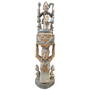 Könnte beinhalten: Eine Holzskulptur einer stehenden Figur mit mehreren Köpfen und Figuren obenauf. Die Skulptur ist in Braun-, Blau- und Weißtönen bemalt.