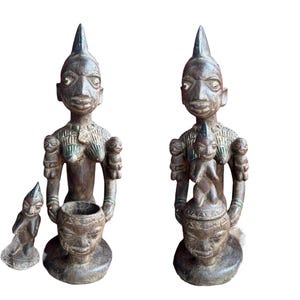 Könnte beinhalten: Bronzefarbene Skulpturen stilisierter menschlicher Figuren. Die zentralen Figuren haben längliche Köpfe und halten Gefäße. Eine Figur hat eine kleine Kinderfigur auf dem Kopf. Eine kleinere Figur kniet in der Nähe. Die Skulpturen haben ein verwittertes Aussehen.