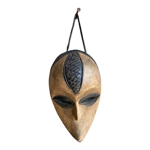 May include: A carved wooden mask with a light brown finish, featuring a dark, textured, diamond-patterned forehead panel and dark, almond-shaped eyes. The mask has a pointed chin and is suspended by a black cord from a metal nail.