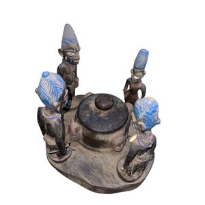 Puede incluir: Escultura de madera tallada con una olla central oscura rodeada de cuatro figuras con detalles azules y negros. Las figuras tienen cabezas alargadas y están adornadas con collares. La base es de forma oscura e irregular.
