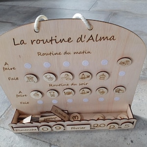 Peut inclure: Un tableau en bois avec un calendrier et un tableau de routine quotidienne pour un enfant nommé Alma. Le tableau comporte un emplacement pour tenir de petits jetons en bois avec des images d'activités comme s'habiller, manger et se brosser les dents. Le calendrier affiche la date du 9 octobre 2025.