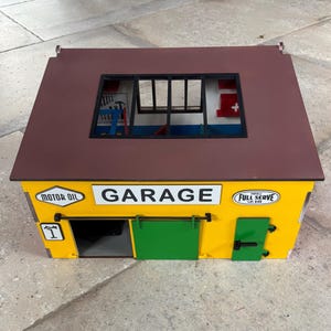 May include: A yellow toy garage with a brown roof and green doors. The word "GARAGE" is displayed in white letters above the green doors. The garage has a window and the words "MOTOR OIL" and "FULL SERVE" are also visible.