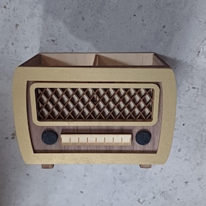 Puede incluir: Organizador de escritorio de madera con forma de radio vintage. El organizador tiene un marco pintado de dorado, una rejilla de altavoz marrón y dos perillas negras. Una escala está impresa debajo de la rejilla. La parte superior tiene dos compartimentos.