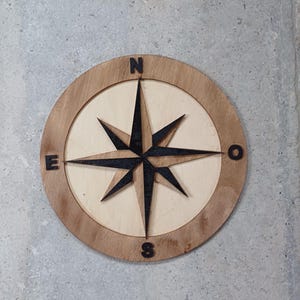 Peut inclure: Une boussole en bois avec une rose des vents noire et les lettres N, E, S et W autour du bord.