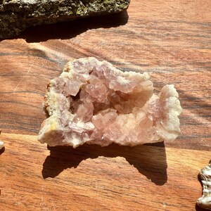 May include: A light pink crystal geode with a hollow centre and a rough, textured exterior. The geode is set on a wooden surface, with a piece of bark in the background. The crystal has a natural, organic appearance.