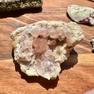 May include: A natural rose quartz geode with a rough, beige exterior and a cluster of pink crystals inside. The geode is displayed on a wooden surface, showcasing its natural beauty and unique formation.