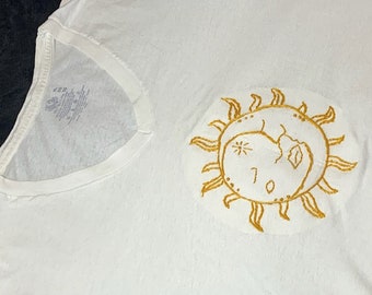 May include: White t-shirt with a gold embroidered sun and moon design.
