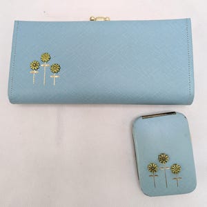 May include: A light blue wallet and matching compact mirror. Both items feature a gold clasp and a floral design. The wallet is rectangular with a clasp closure. The compact mirror is small and rectangular.