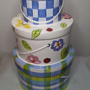 May include: A set of three ceramic nesting boxes with a floral and checkered design. The top box is blue and white checkered, the middle box is white with flowers and ladybugs, and the bottom box is green, blue, and white checkered.