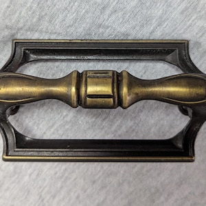 May include: An antique-style drawer pull with a brass-colored handle and a dark bronze-colored frame. The handle has a cylindrical center and conical ends. The frame has a rectangular shape with decorative cutouts.