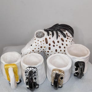 May include: A set of four ceramic mugs with animal-shaped handles. The mugs are white with black and brown spots. The handles are shaped like a horse's head, a cow's head, a pig's head, and a sheep's head.