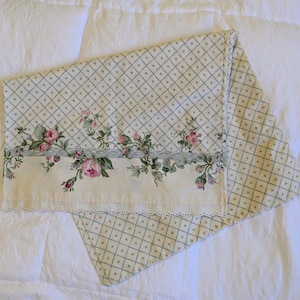 May include: A rectangular pillowcase featuring a cream-colored background with a light green diamond grid pattern. A horizontal border displays pink roses with green leaves and vines, finished with a white scalloped eyelet lace trim edge.