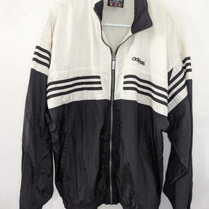 May include: A vintage Adidas track jacket in black and cream. The jacket features the Adidas logo on the chest and three black stripes on the sleeves. It has a full zipper and a stand-up collar. The jacket is hanging on a white hanger.