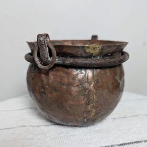 May include: A small, antique copper cauldron with a rounded body and a wide rim. The cauldron has a dark, aged patina with visible wear and tear. A metal ring handle is attached to the side. The cauldron is likely used for decorative purposes.