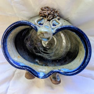 May include: A handmade ceramic bowl with a unique face design. The bowl features a dark blue rim and a light blue and gray interior. The face has blue eyes and a nose, with a textured brown hair-like detail on top. The bowl rests on three small feet.