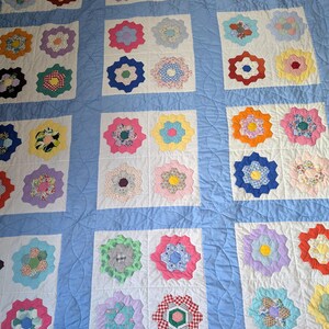 May include: A quilt featuring a light blue background with white square blocks. Each white block contains a multi-colored floral-patterned hexagon flower motif. The fabric is stitched with a circular quilting pattern throughout the blue sections.