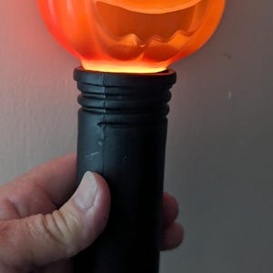 May include: A handheld light-up toy featuring a bright orange plastic jack-o'-lantern head with a carved face and a black cylindrical handle. The pumpkin glows from within, illuminating the molded eyes, nose, and wide, jagged, smiling mouth.